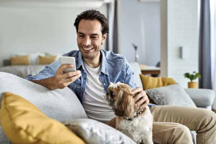 Happy man texting on mobile phone while relaxing with his dog at home.